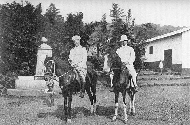 Das Bild zeigt zwei Kolonialoffiziere in Uniform, die auf ihren Pferden sitzen, die vor dem Bismarck Brunnen in Kamerun stehen. Sie blicken in die Kamera. Hans Dominik (rechts) war ein Kolonialoffizier der Schutztruppe, der als dienstältester Offizier in der Kolonie Kamerun ein brutales und mörderisches Erbe hinterließ. Im Sammlerwiki wird auch angegeben, dass er (wahrscheinlich geplünderte) Gegenstände in die Sammlungen des Linden-Museums und des Ethnologischen Museums Berlin eingebracht hat.