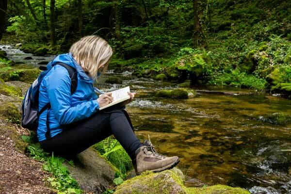 A person sitting next to a stream, writing in a book