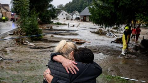 Two women hug each other after being evacuated by rescue teams in Jesenik, Czech Republic on 15 October, 2024. Emergency workers in protective gear stand in front of damaged houses and large amounts of debris in brown flood waters