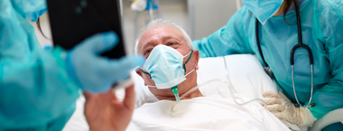 A patient lying in a hospital bed wearing a face mask and surrounded by gowned hospital staff and using an ipad