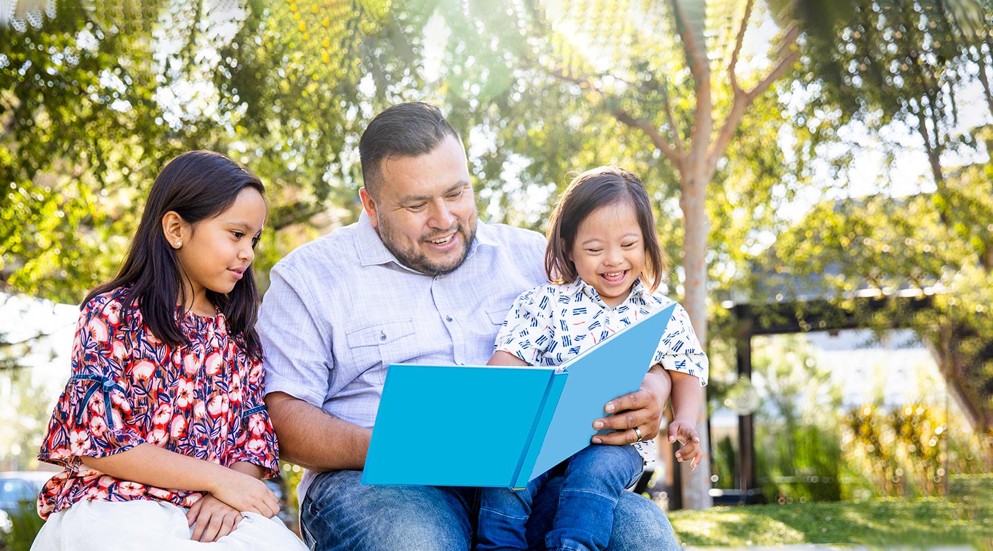 Dad sits outside with an open book; he reads to his two young children. 