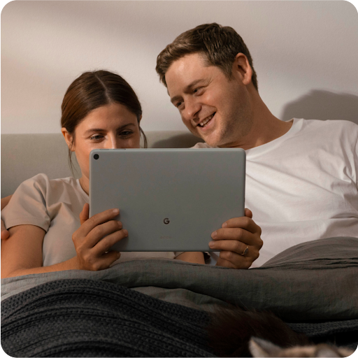 Duas pessoas relaxando na cama e sorrindo enquanto assistem conteúdo no tablet.