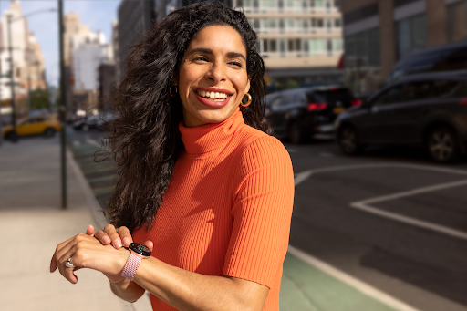 The smartwatch user is standing on the sidewalk in a downtown city area and smiling while looking in the direction that the Google Maps watch is navigating toward.