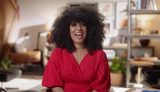 A woman, wearing a red top, laughs while facing the camera. An office space is behind her.