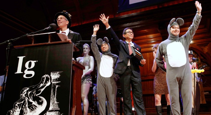 A man stands at a lectern that reads "Ig", while two men dressed as mice and a man in a suit wave to the crowd