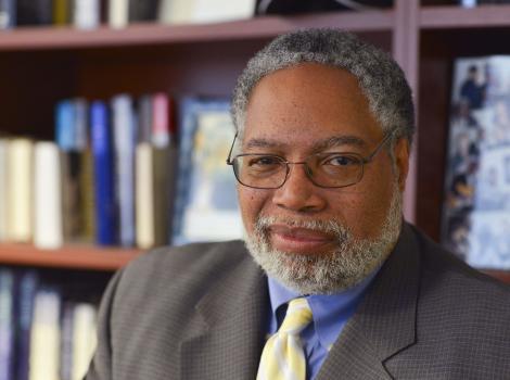 Gray haired man with a beard seated in front of a bookcase