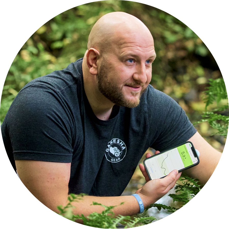 A man kneels in the undergrowth of a forest, referring to graphic data displayed on two smartphone screens.