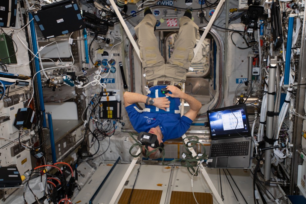 An astronaut floats upside down in the microgravity environment of the International Space Station, wearing a virtual reality headset.