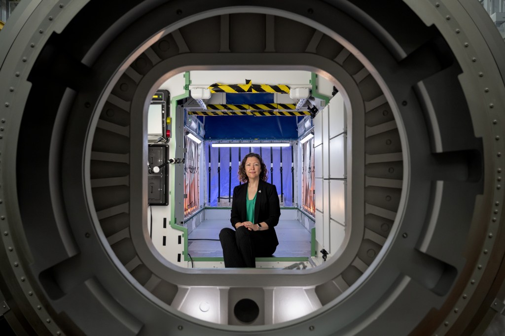 Stephanie Dudley, Gateway's mission integration and utilization manager, sits inside a high-fidelity mockup of HALO (Habitation and Logistics Outpost) at NASA’s Johnson Space Center. She is framed by a docking port, with interior components visible behind her.The mockup’s design showcases the compact and functional environment Artemis astronauts will use for living, conducting experiments, and preparing for lunar surface missions. Photo credit: NASA/Josh Valcarcel