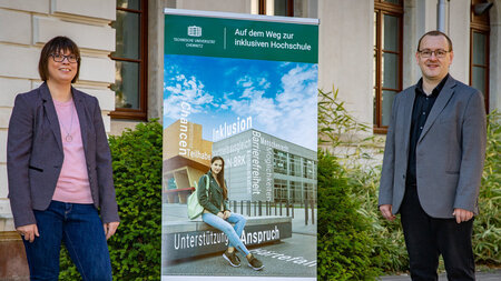 EIne Frau und ein Mann in Blazer und Sakko stehen neben einem Schild. Darauf steht Inkluaisve Hochschule.